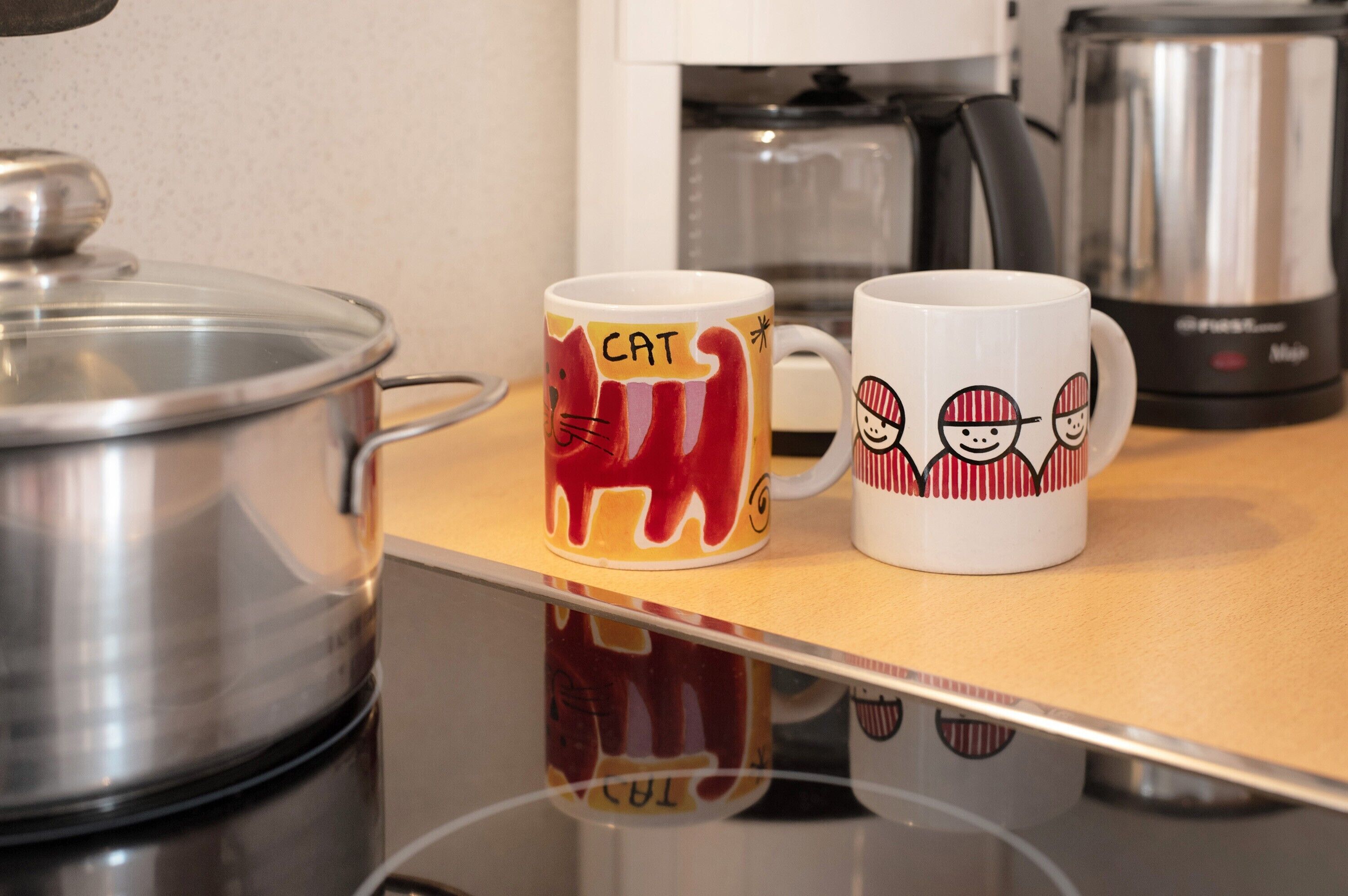 Två kaffemuggar står på en köksbänk, med en kastrull och en kaffebryggare i bakgrunden.