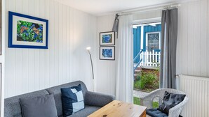 Huoneessa on harmaa sohva, puinen pöytä ja rottinkituoli. Seinällä on kehystettyjä kuvia, ja ikkunasta näkyy sininen rakennus.