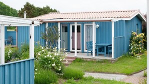 Un bâtiment en bois bleu, doté d’un patio couvert, d’une porte et d’un petit jardin agrémenté de fleurs et d’une chaise.