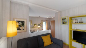 Un salon avec un canapé, un téléviseur posé sur un meuble jaune, une lampe et une fenêtre avec des rideaux.