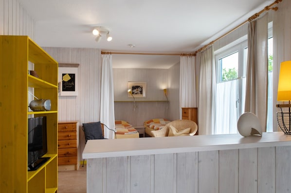A room with a white wall, a wooden bookshelf, a bed with a patterned bedspread, a wicker chair, and a window with curtains.