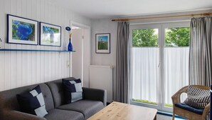 A living room with a sofa, a chair, a table, and a window with curtains. - Holiday home "Möve ABA Bungalow 63" with private terrace and WLAN (Rerik)