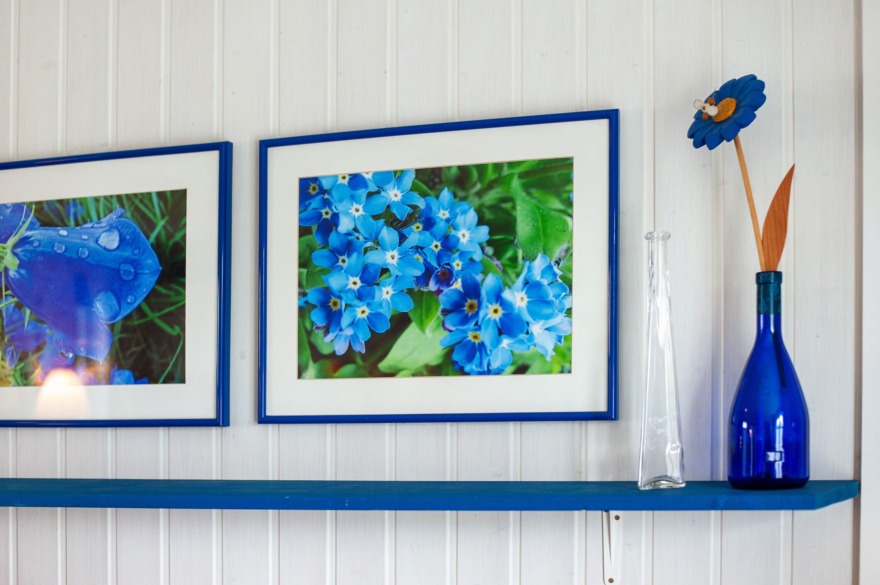 Two framed pictures of blue flowers on a white wall, a clear glass vase with a blue bottle, and a blue vase with a flower on a shelf.