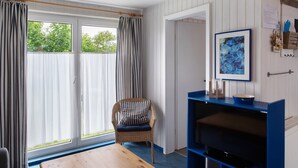 A room with a wicker chair, a wooden table, a blue entertainment unit, and a large window with curtains. - Holiday home "Möve ABA Bungalow 63" with private terrace and WLAN (Rerik)