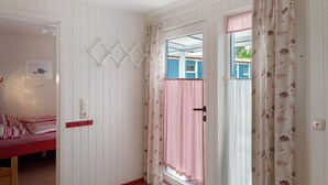 A room with a bed, a door with a light switch, and a window with a view of a blue building. - Holiday home "Möve ABA Bungalow 62" with private terrace and WLAN (Rerik)
