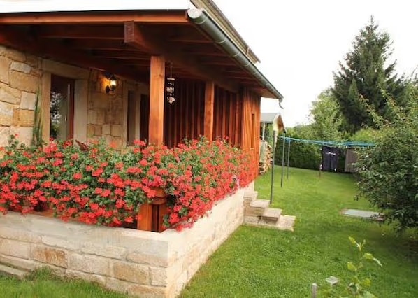 Un bâtiment en pierre avec un porche en bois, un parterre fleuri avec des fleurs rouges et une pelouse verte.