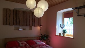 A bedroom with a wooden ceiling, a bed with red bedding, a window with a heart-shaped decoration, and a hanging light fixture.
