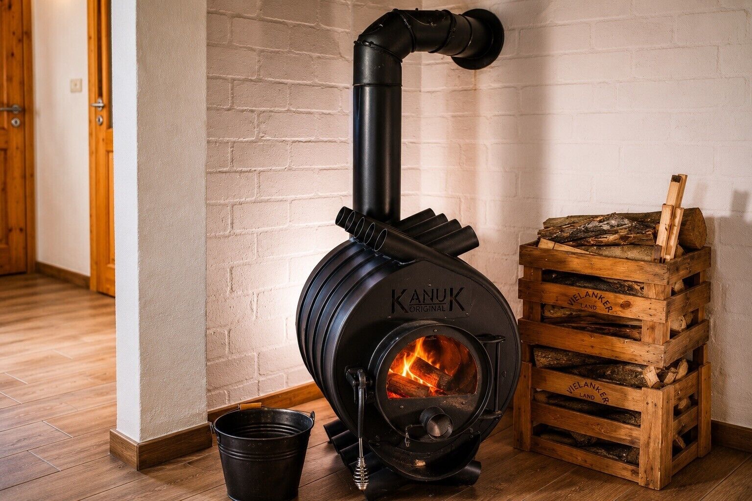 Un poêle à bois noir avec un feu visible, une palette en bois remplie de bûches et un seau en métal.