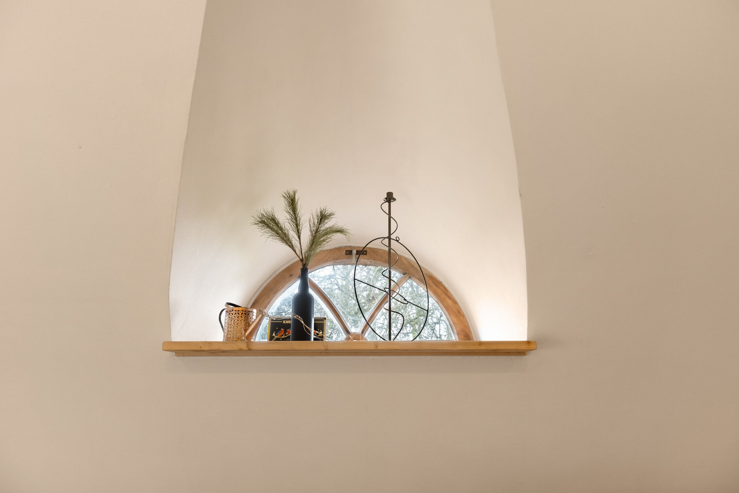 Una ventana con un estante de madera, un florero con plantas y un adorno metálico.