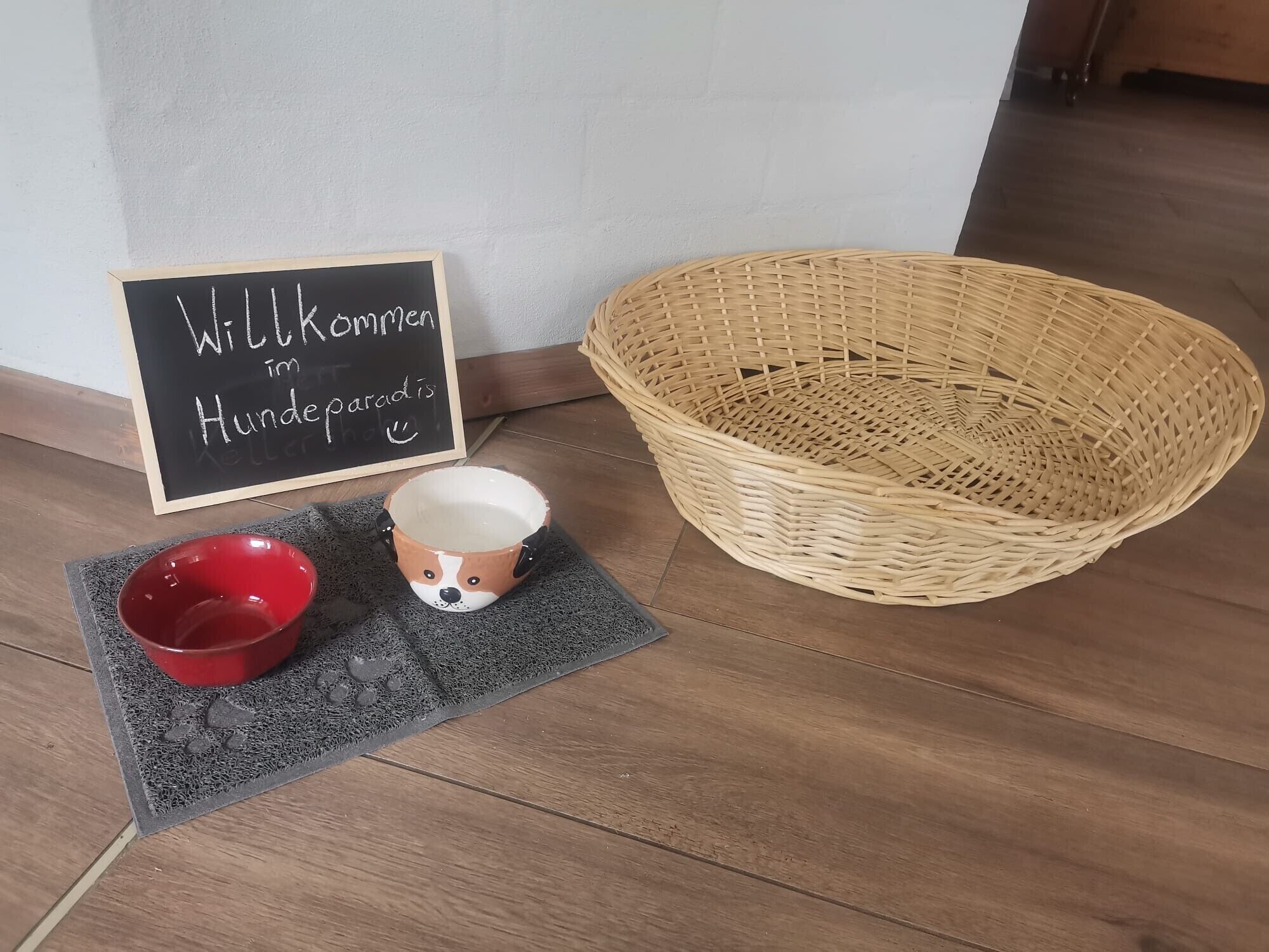 Un panier en osier, un bol rouge, une tasse en céramique ornée d’un visage de chien et un tapis d’accueil.