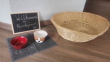 Un panier en osier, un bol rouge, une tasse en céramique ornée d’un visage de chien et un tapis d’accueil.