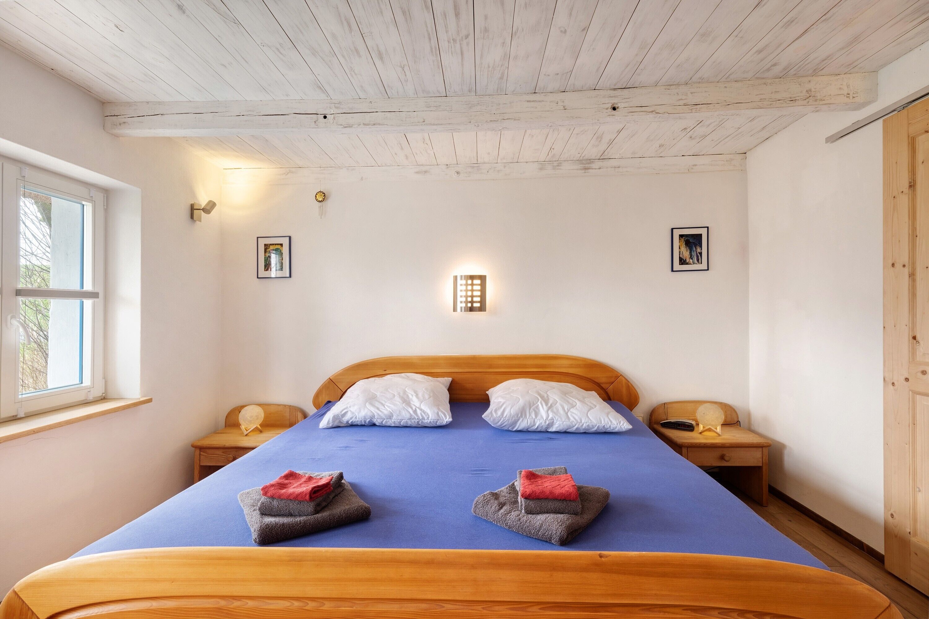 Une pièce avec un plafond en bois, un lit avec une literie bleue, deux tables de chevet et une fenêtre.