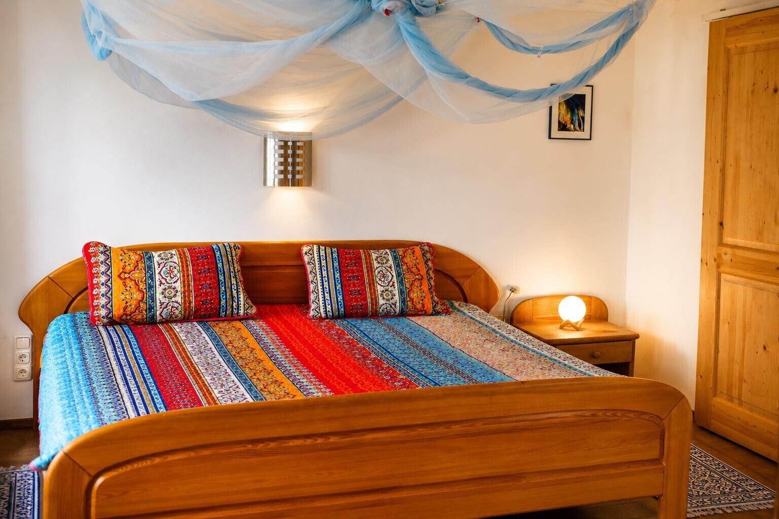 A bed with a colorful blanket and pillows, a bedside lamp, a wall-mounted light, and a wooden wardrobe.