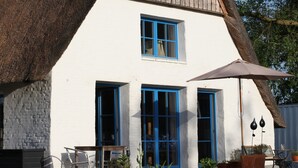 Ein Haus mit einem Strohdach, weißer Fassade und blauen Fensterrahmen, mit einer kleinen Sitzgelegenheit mit Stühlen und Tischen.