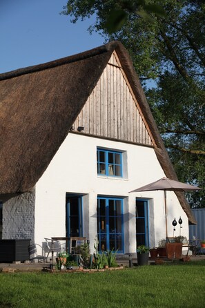 Une maison à toit de chaume, aux murs blancs et aux fenêtres à cadre bleu, avec un petit espace extérieur aménagé avec des chaises et des tables.
