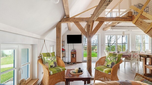 Ein geräumiges Zimmer mit hoher Decke, Holzbalken, Korbmöbeln, einem Fernseher und großen Fenstern.