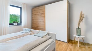 Un dormitorio con cama, ventana con cortinas, pared de paneles de madera, armario blanco y una mesita con un jarrón.