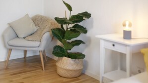Una habitación con un sillón de color claro, una maceta tejida, una mesita de noche con lámpara y suelo de madera.
