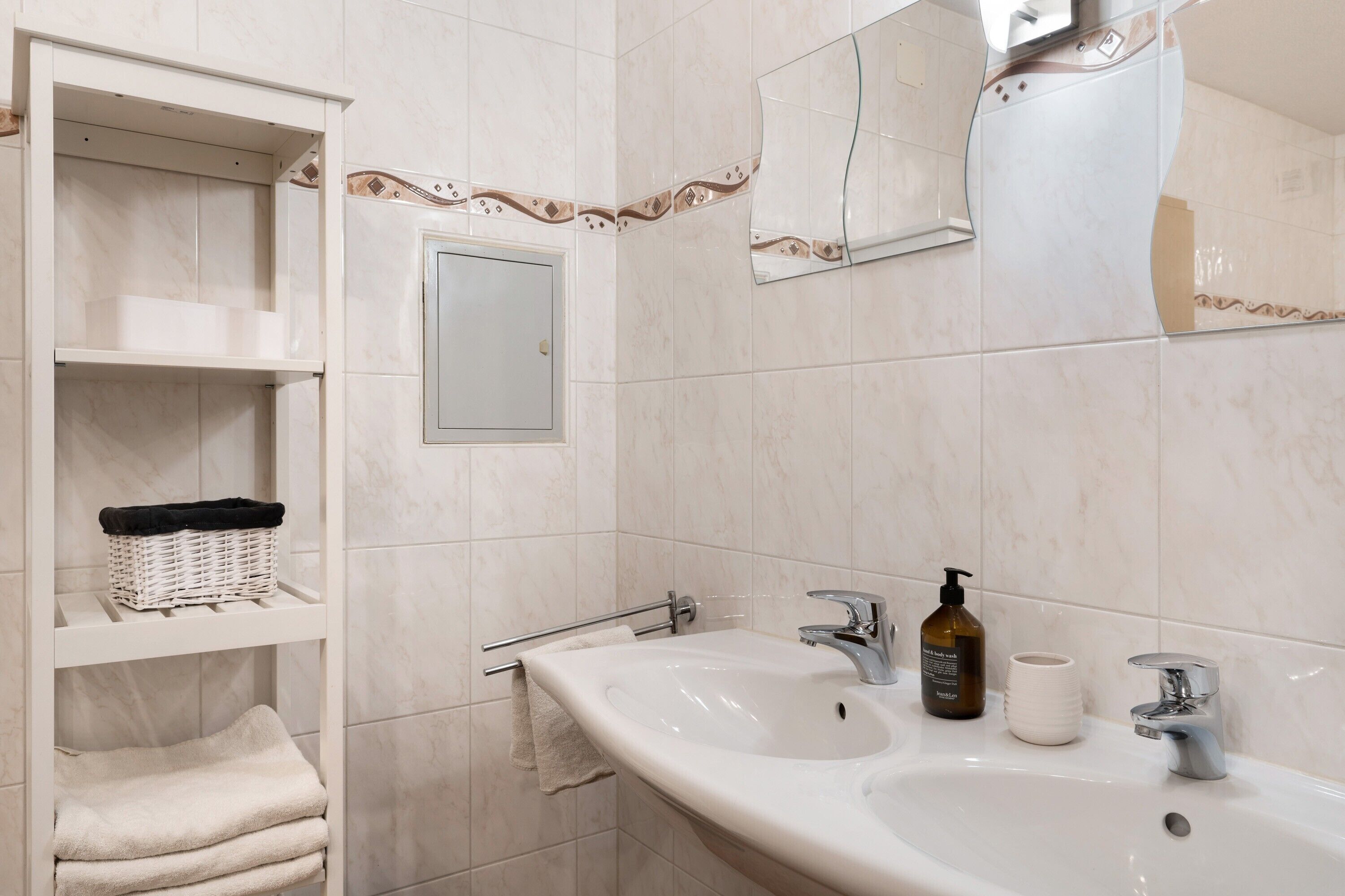 Ein Badezimmer mit zwei Waschbecken, einem Spiegel, einem Handtuchhalter und einem Regal mit Korb und Handtüchern.