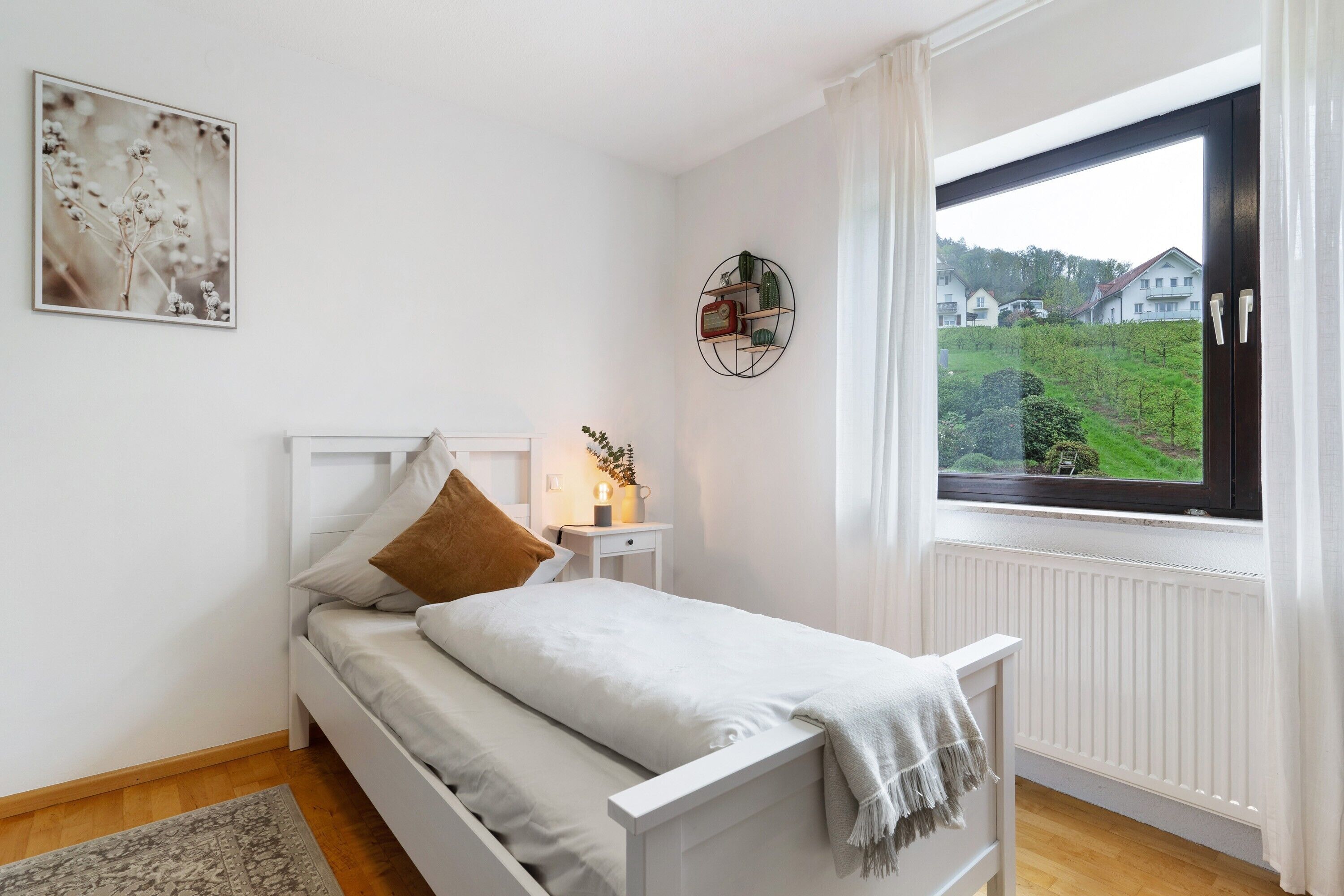 Ein Schlafzimmer mit einem Bett, einem Nachttisch, einem Regal an der Wand und einem Fenster mit Blick auf Häuser.