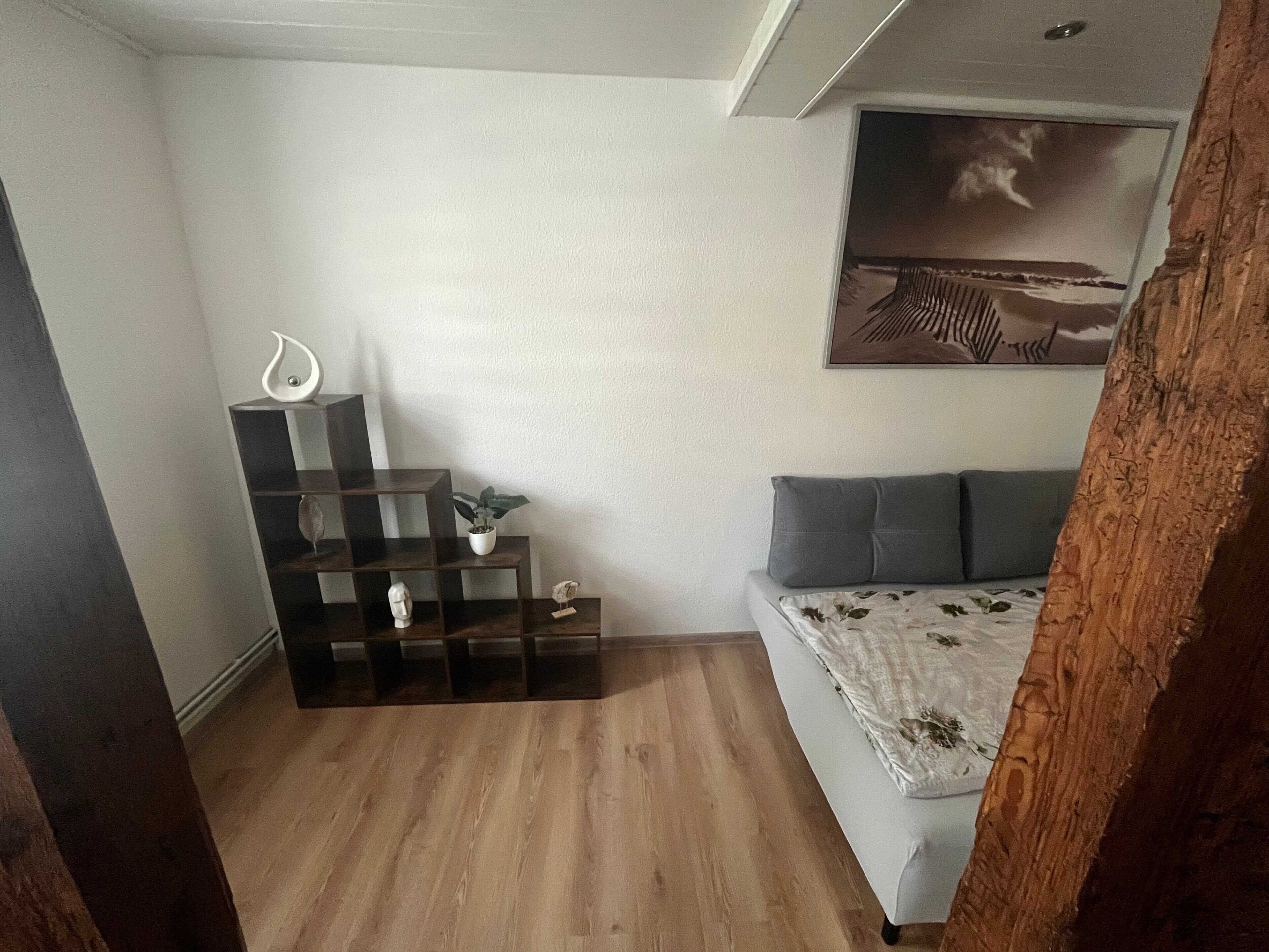 A room with a bed, a wooden floor, a white wall, a shelf with decorative items, and a painting.