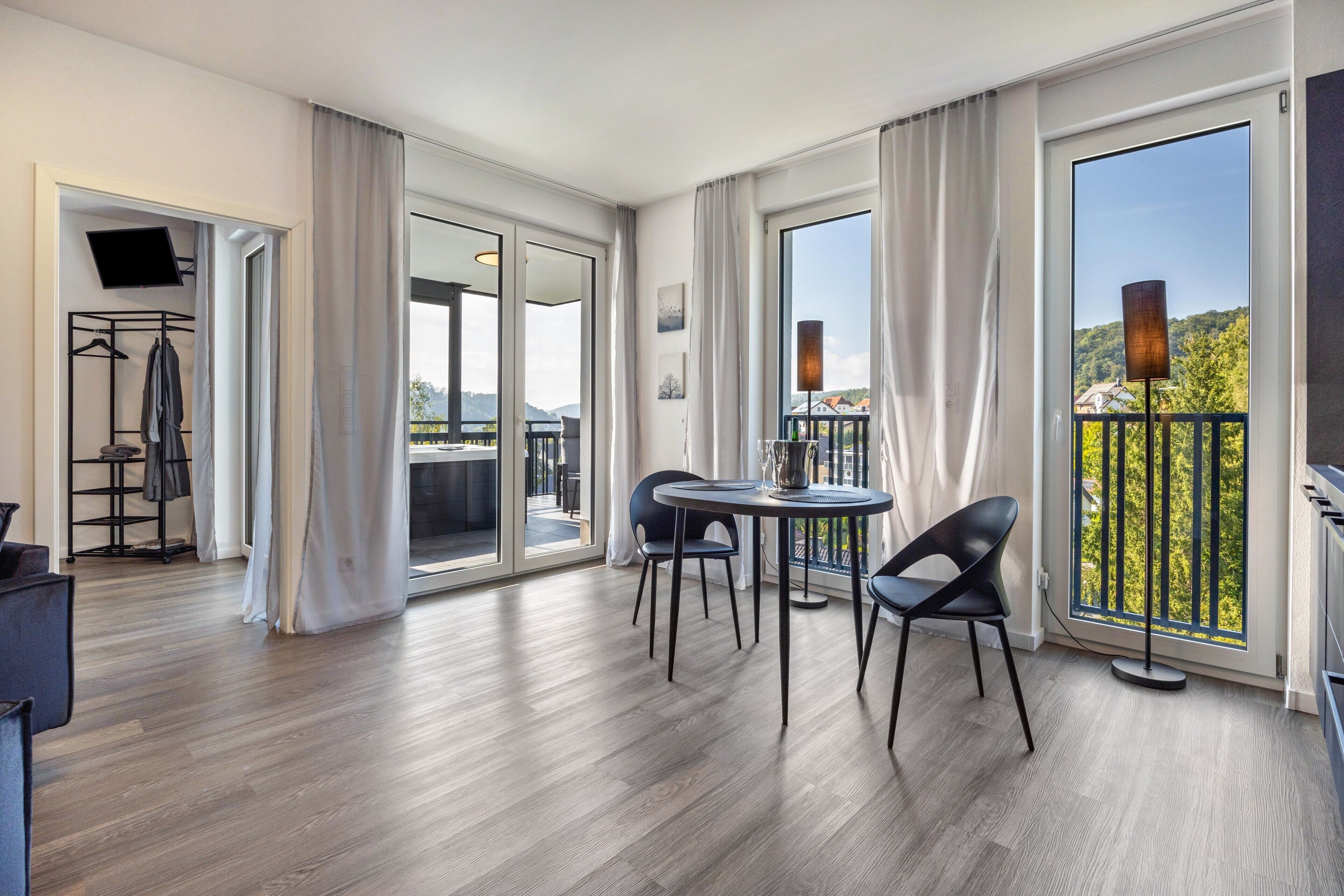Una habitación de hotel moderna con balcón, zona de comedor con sillas, televisor de pantalla plana y perchero.