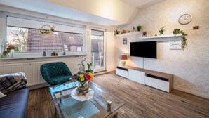 A modern living room with a sofa, a coffee table with a vase, a TV mounted on the wall, and a window with a view of the outside. - Apartment 'Haus Heidorn' with Balcony and Wi-Fi (Sittensen)