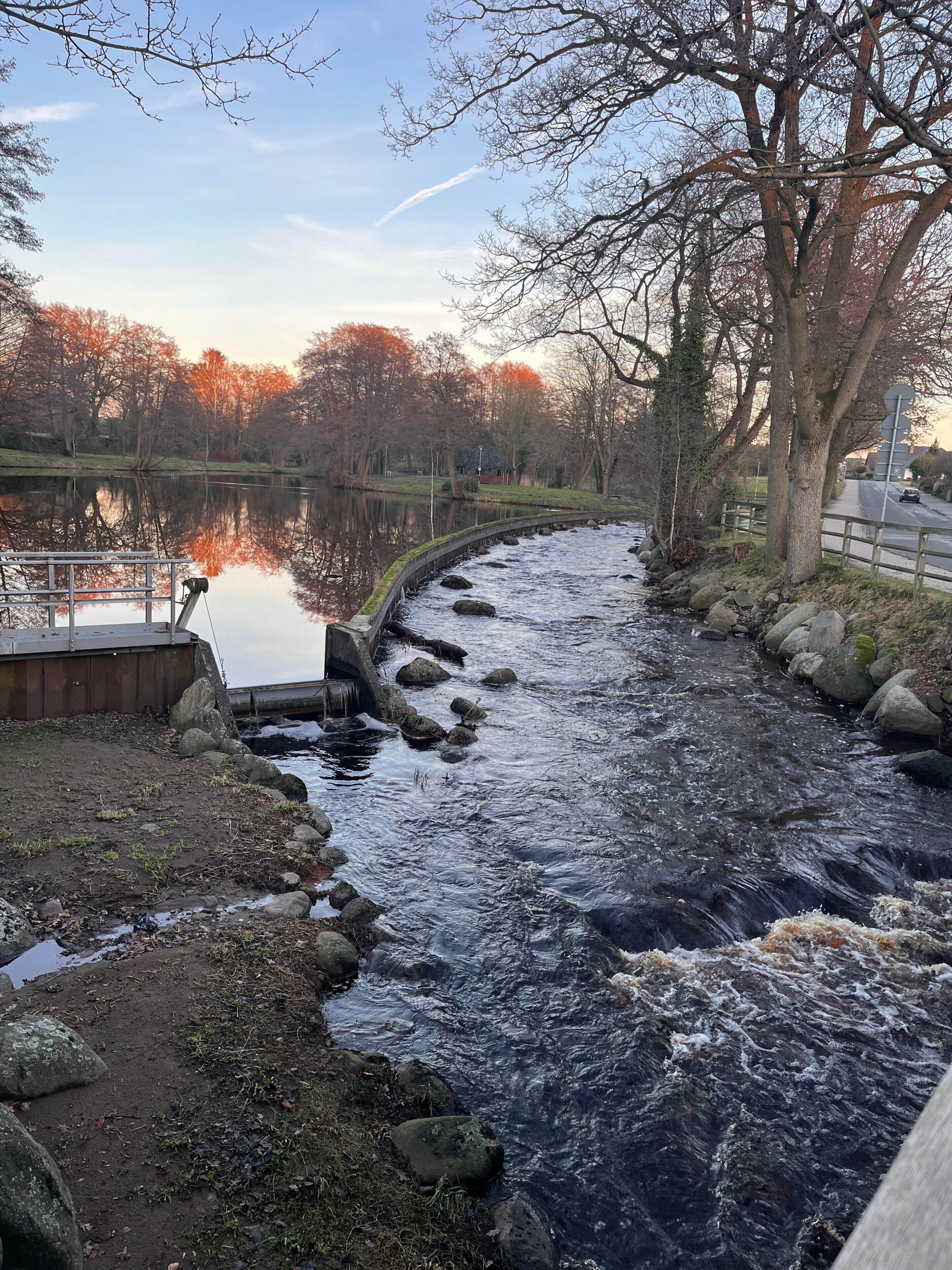En flod der løber gennem en park med en bro, træer og et roligt vandareal, der spejler himlen.