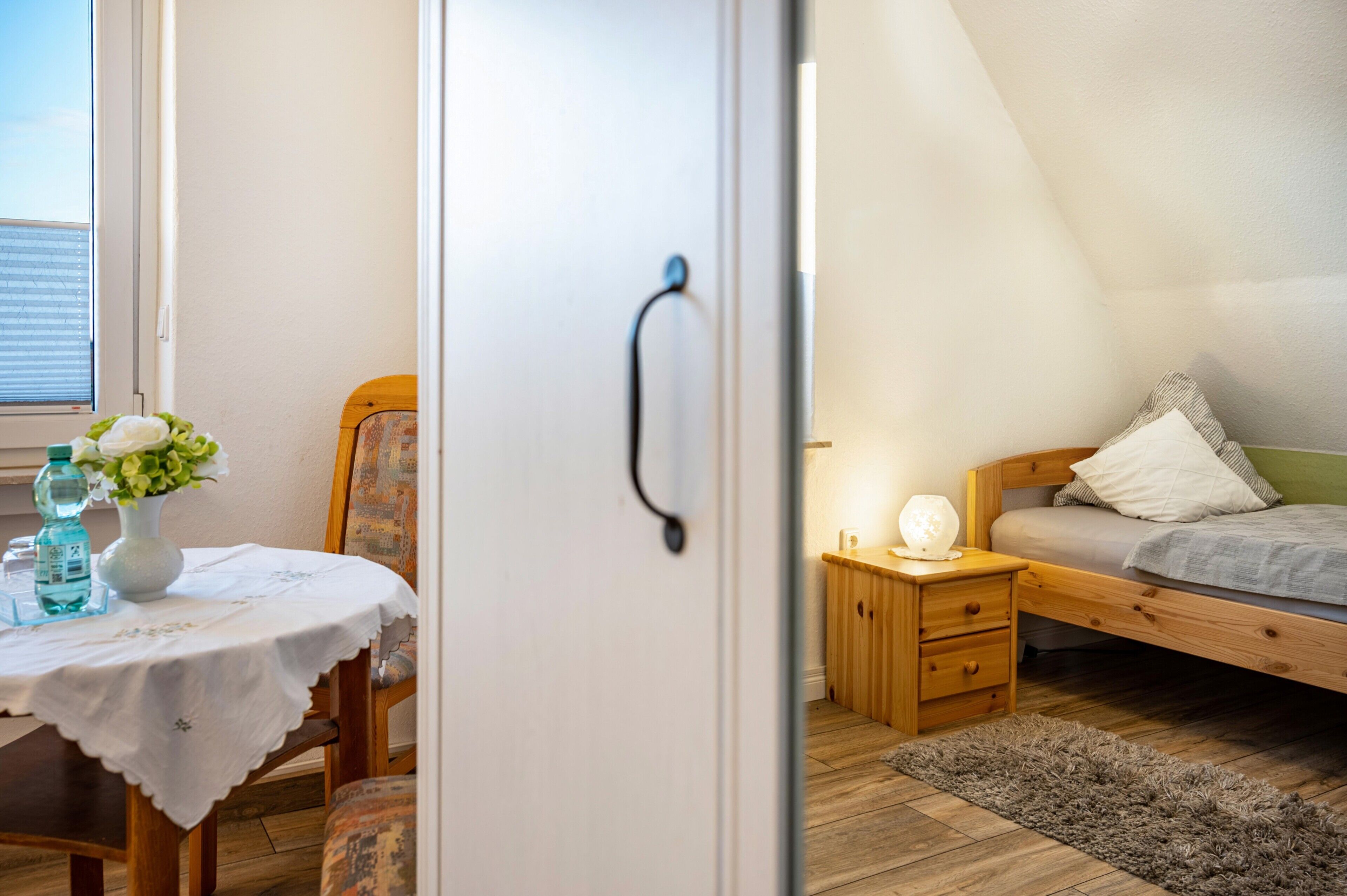 A bedroom with a wooden bed, a bedside table, a nightstand, a small table with a vase of flowers, and a chair.