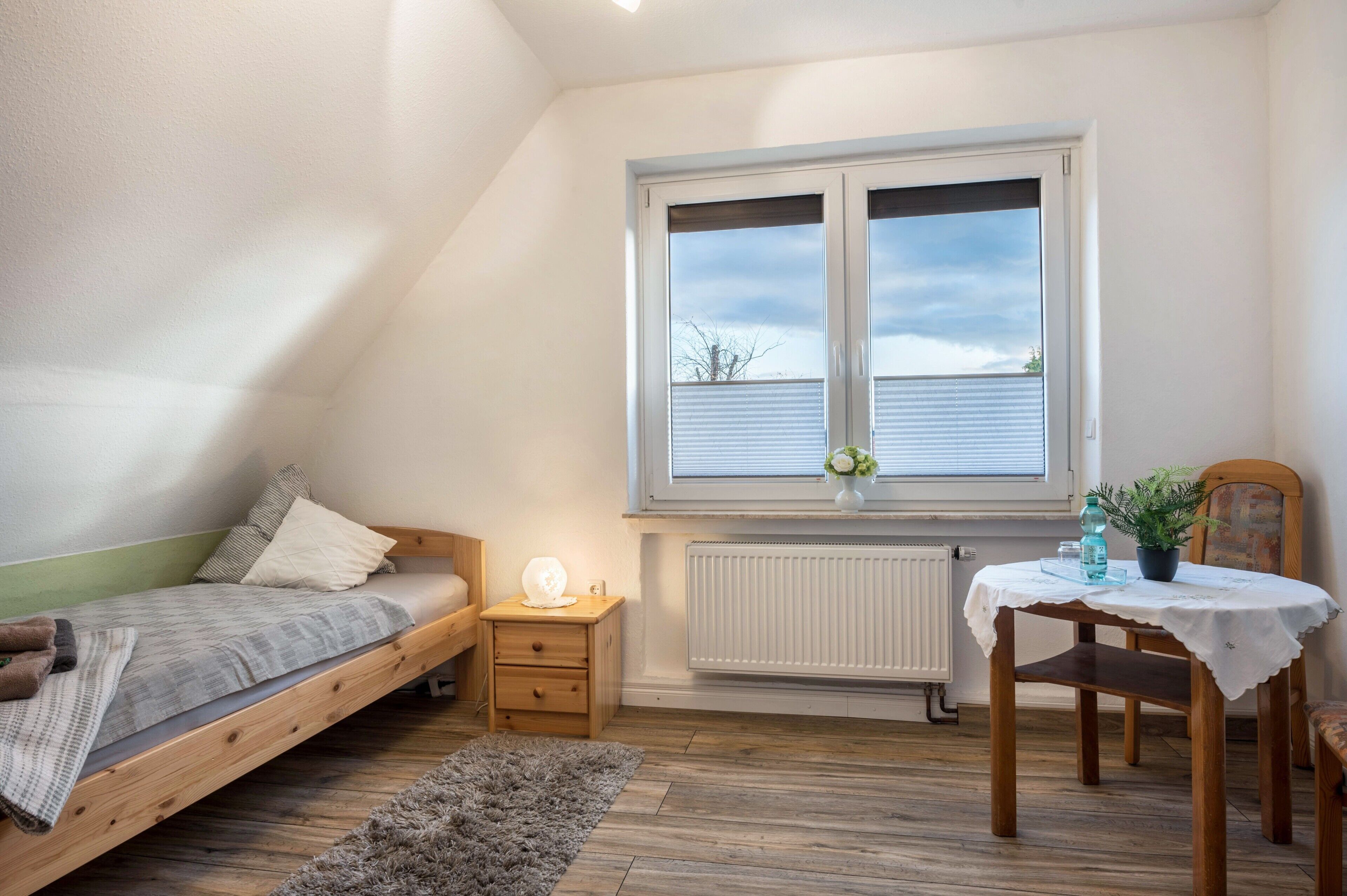Een slaapkamer met een enkel bed, een klein nachtkastje, een stoel, een tafel met een vaas en een raam met jaloezieën.