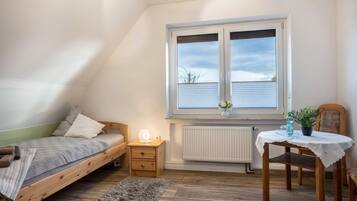 Een slaapkamer met een enkel bed, een klein nachtkastje, een stoel, een tafel met een vaas en een raam met jaloezieën.