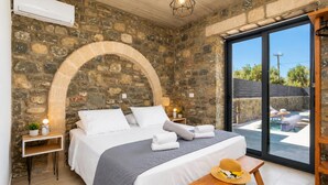 Une chambre aux murs de pierre, comprenant un lit, des tables de chevet et donnant sur un espace extérieur avec piscine.