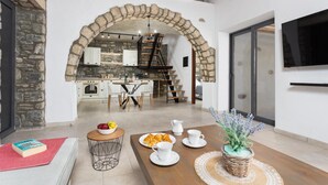 Un intérieur moderne avec une arche en pierre menant à la cuisine, un coin repas avec une table mise pour le petit-déjeuner et un espace de vie avec un téléviseur et une plante.