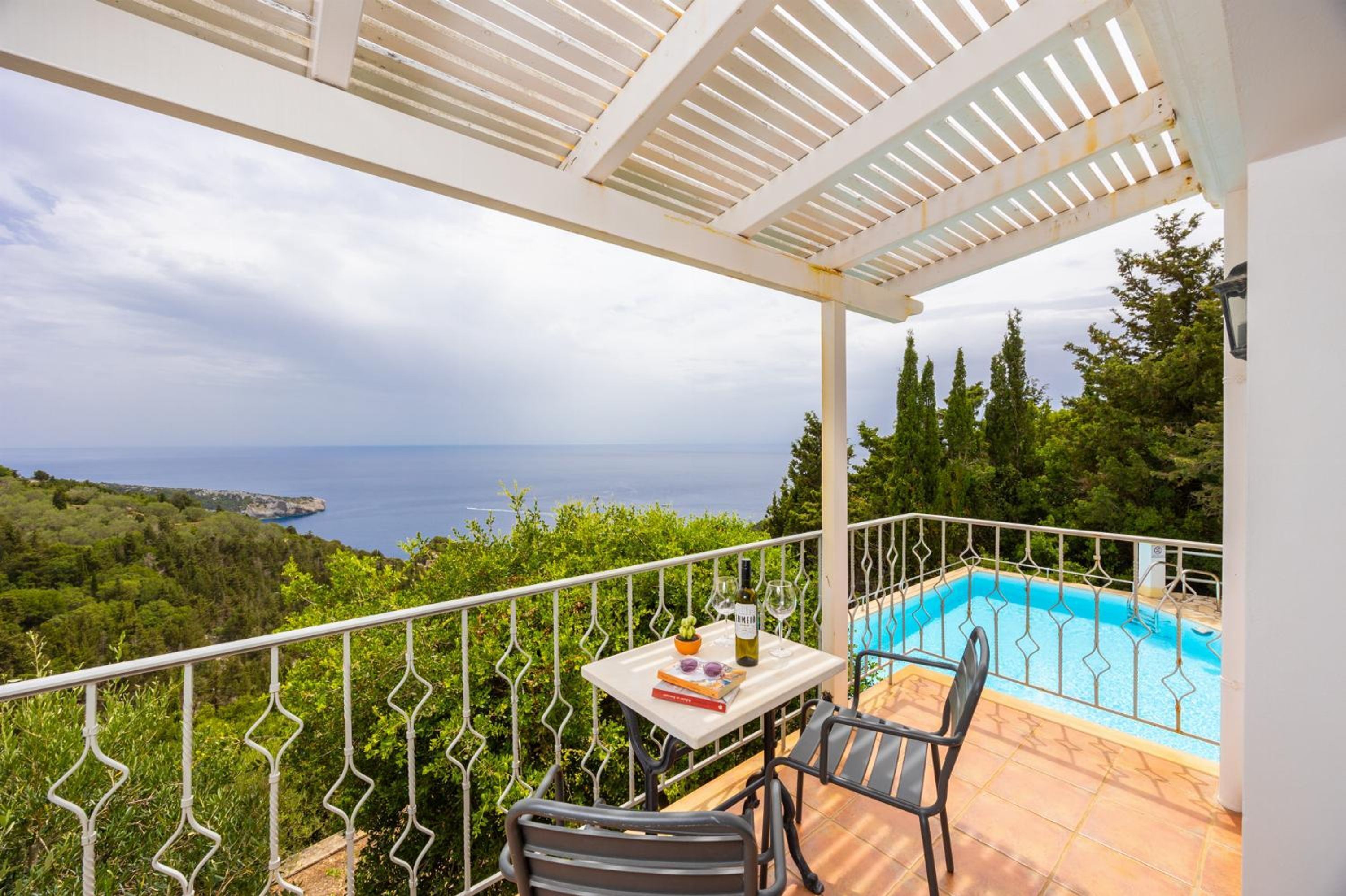 A balcony with a pool, a table with a book, a bottle, and a glass, overlooking a coastal landscape.