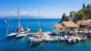A marina with various boats docked, a building with a tiled roof, and a clear blue sky. - Villa Gardenia in Paxi (Paxos)