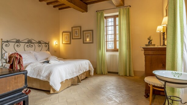 A bedroom with a double bed, a bedside table, a dresser, a window with curtains, and a small table with a lamp. - Il Poggio Alle Trese 14 Emma Villas (Citta della Pieve)