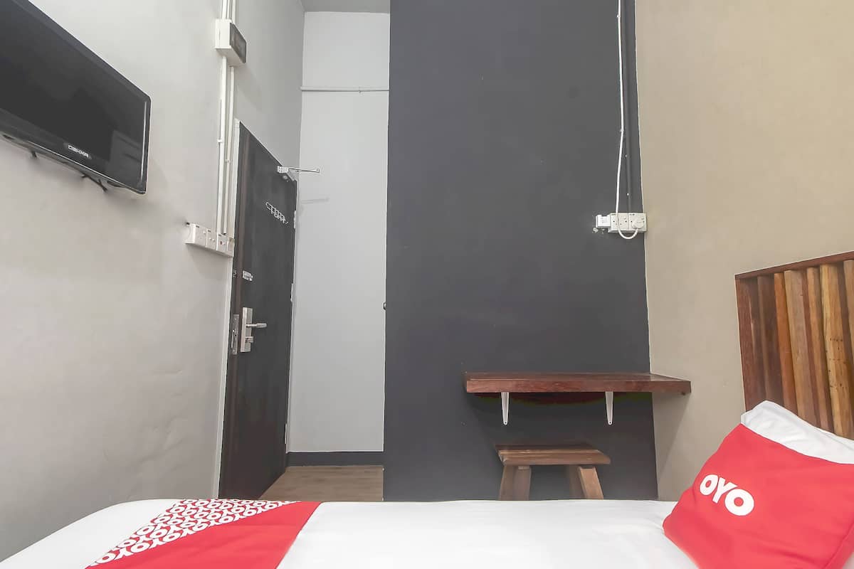 A small, compact room with a bed, a TV mounted on the wall, a wooden bench, and a door leading to another room.