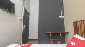 A small, compact room with a bed, a TV mounted on the wall, a wooden bench, and a door leading to another room.