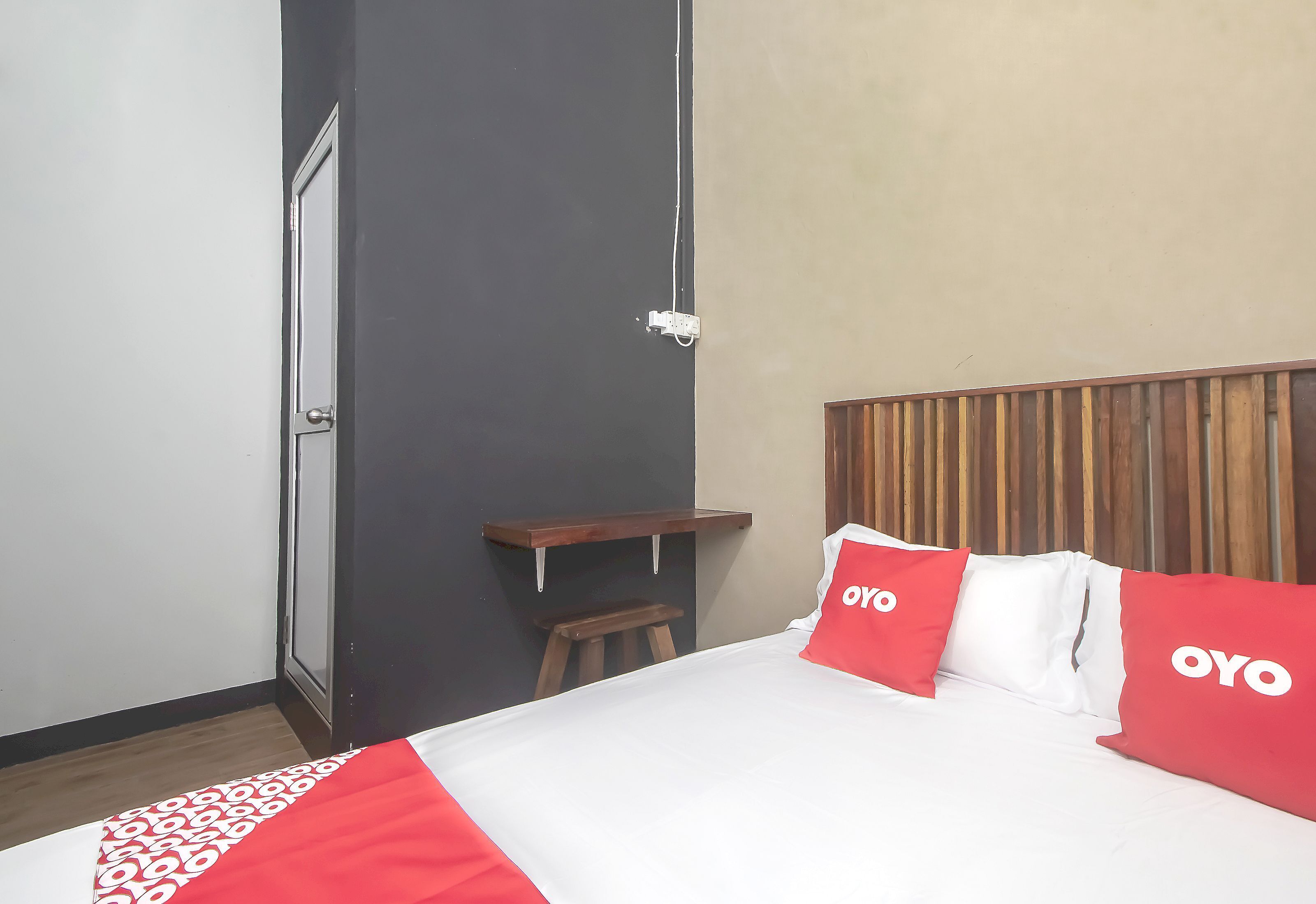 Habitación de hotel con cama, cabecero de madera, taburete pequeño de madera, estante y toma de corriente en la pared.