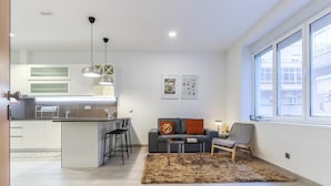 A modern kitchen with a central island, bar stools, pendant lighting, and a grey sofa with a brown rug.