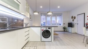 A modern kitchen with a built-in washing machine, pendant lights, and a view of the street through large windows.