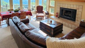 Ein geräumiges Wohnzimmer mit einer großen Eckcouch, einem Kamin und einem Fernseher. Mehrere Fenster sorgen für natürliches Licht und Ausblicke ins Freie.