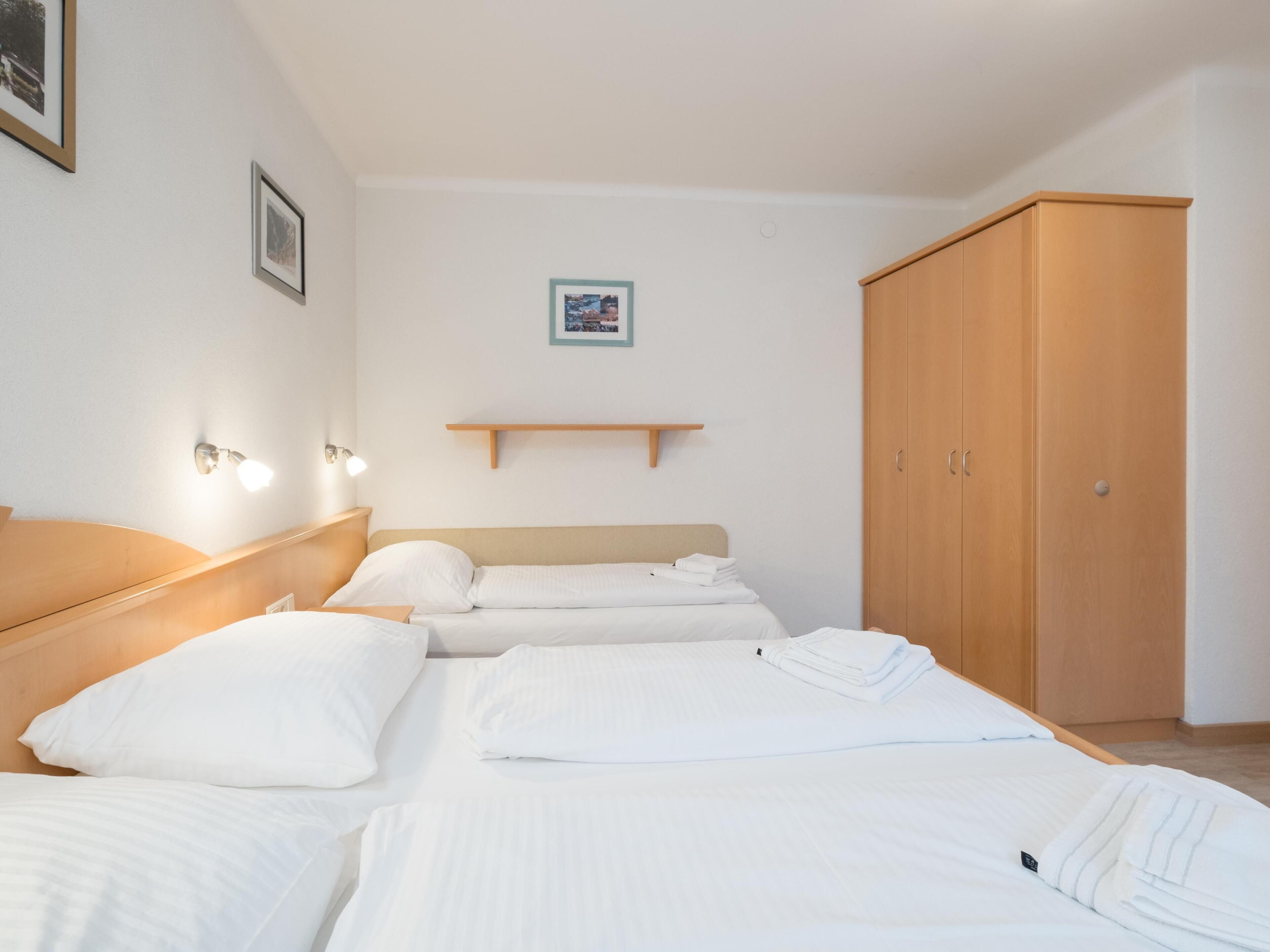 Une chambre d’hôtel avec deux lits, une armoire en bois, une petite étagère et des lampes fixées au mur.