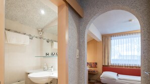 Une salle de bain équipée d’un lavabo, d’un miroir et d’une douche. La pièce adjacente est dotée d’une fenêtre avec des rideaux et d’un lit avec une tête de lit rouge.