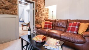 A cozy living room with a stone wall, a brown leather sofa, and a coffee table with a wine bottle and magazines. - The Brewhouse (Church Stretton)