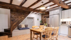 A dining area with a wooden table and chairs, a stone wall, and a kitchen with white cabinets and a microwave. - The Brewhouse (Church Stretton)