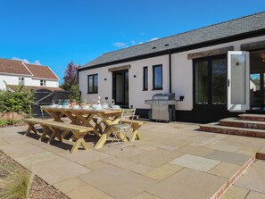 Eine Terrasse mit einem Holztisch, gedeckt für das Essen, einem Grill und einem weißen Gebäude mit schwarzen Fensterrahmen.