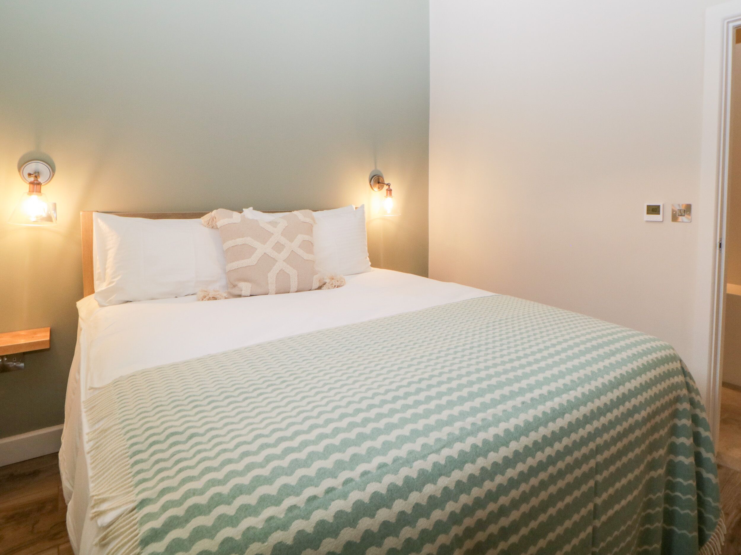 A neatly made bed with a patterned bedspread, two wall-mounted light fixtures, and a door to the right.