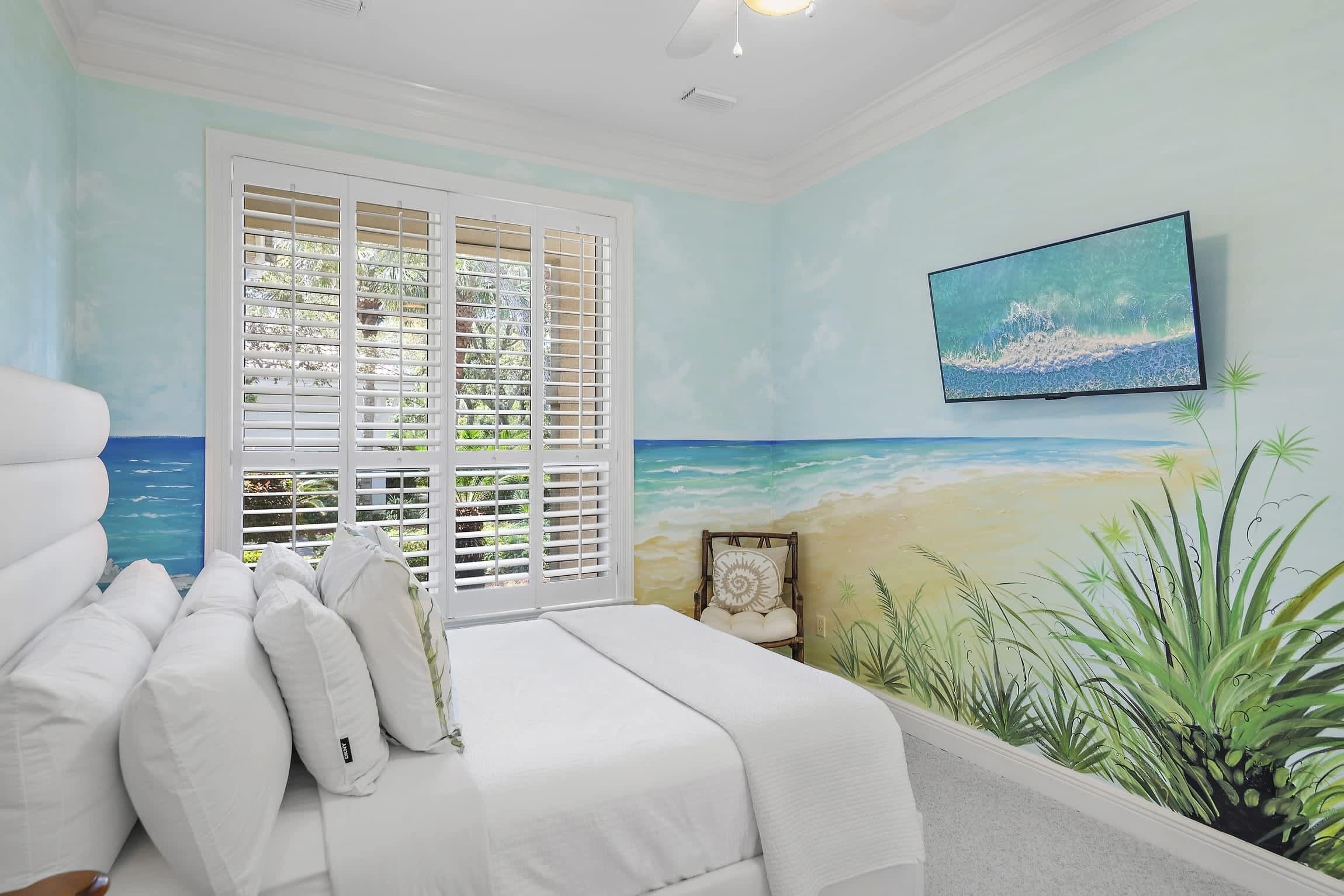 A bedroom with a bed, white bedding, a beach mural, and a television on the wall.