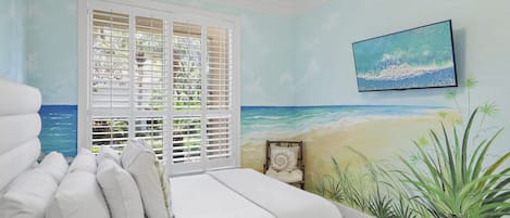 A bedroom with a bed, white bedding, a beach mural, and a television on the wall.
